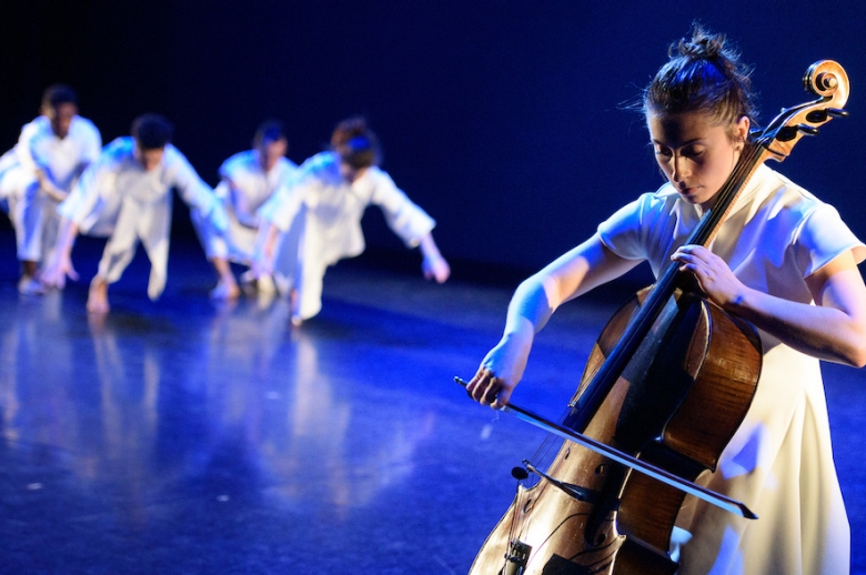 Group of dancers on stage, lit by a blue light, with cellist accompanying them in the foreground